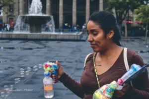 Mujer sostiene pistola de burbujas y botellas mientras trabaja en plaza pública, ejemplo de la economía popular en Colombia.