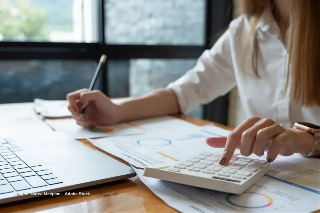 El cuidado frena los micronegocios de las mujeres en Colombia: venden 40% menos 2 Mujer escribe y usa calculadora sobre escritorio con gráficos financieros, ejemplo de emprendimientos femeninos en Colombia.