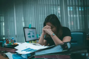 Mujer en oficina con manos sobre el rostro frente a documentos y computadora, reflejo de las dificultades en los micronegocios de mujeres en Colombia.