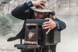 Un hombre con sombrero y traje de época ajusta una cámara antigua de fuelle sobre un trípode en un paraje desértico, en una escena que representa la producción del cine colombiano en la película "Adiós al amigo".