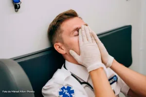 Paramédico con uniforme y guantes sentado, se cubre el rostro con las manos, mostrando agotamiento laboral.