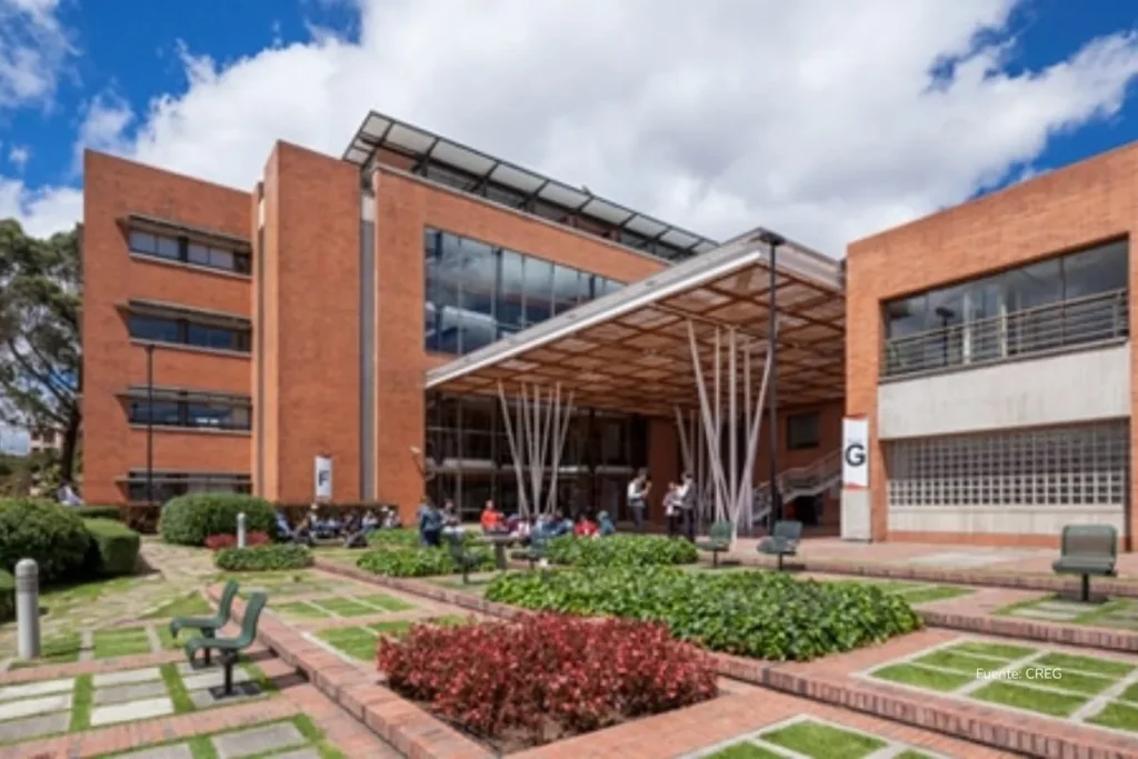 Edificio moderno de ladrillo y vidrio con jardines exteriores, bancos y personas reunidas en la entrada.