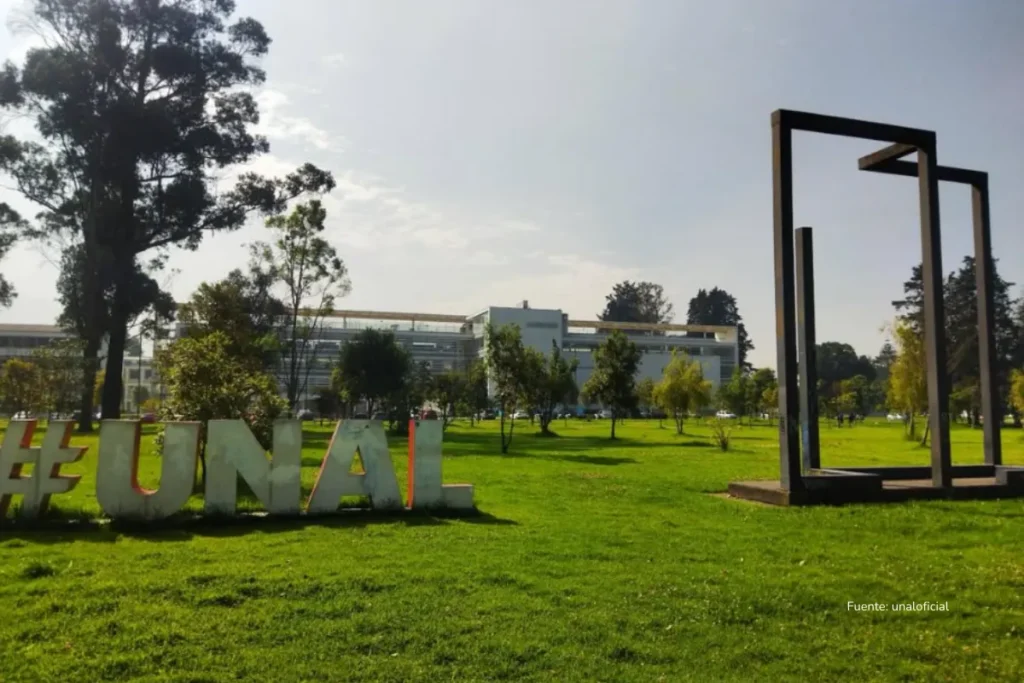 Vista del campus de la Universidad Nacional de Colombia con zonas verdes, la escultura cuadrada metálica y el letrero #UNAL en primer plano.