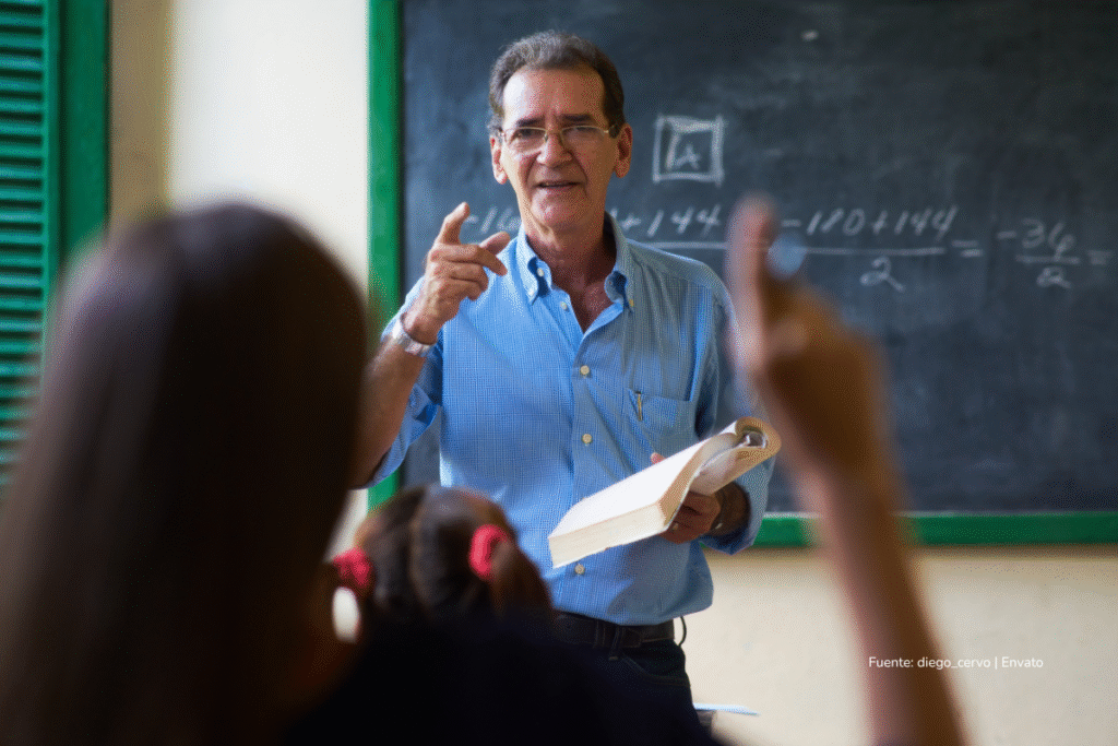 Profesor sosteniendo un libro mientras explica una lección frente a un tablero en el aula, con estudiantes participando y levantando la mano. Aumento salarial docentes 2026