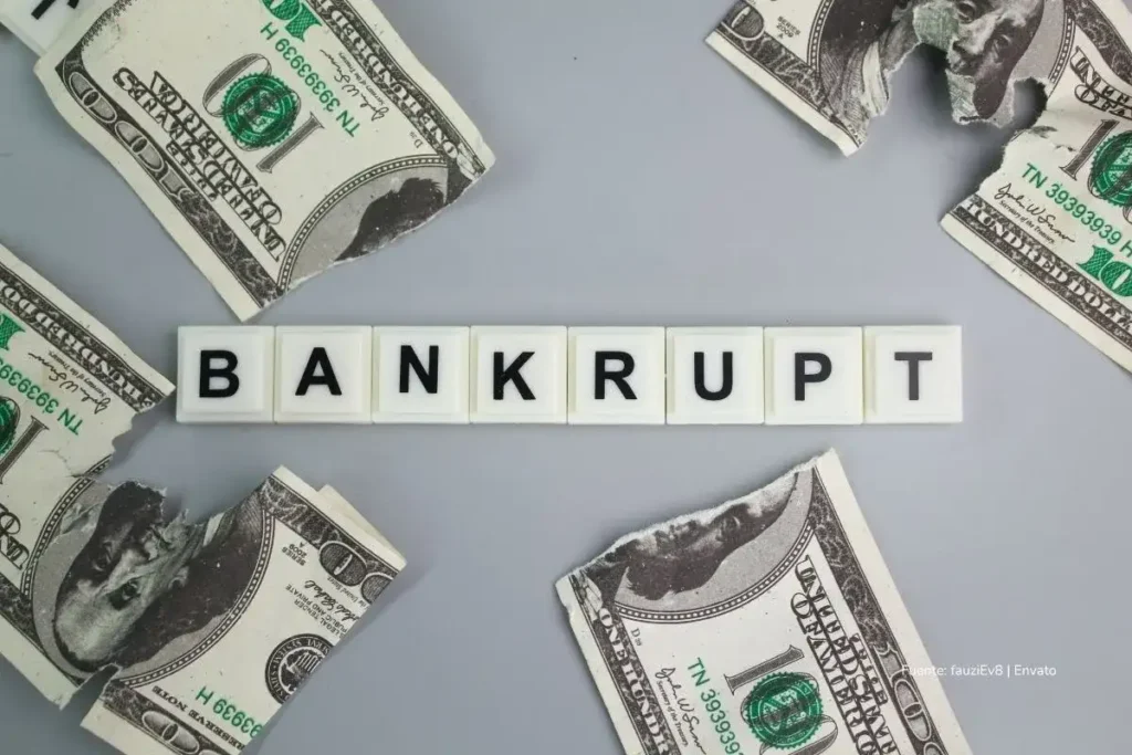 Bancos de Estados Unidos en líos por fraudes, quiebras y pérdidas millonarias que sacuden a Wall Street 2 Billetes de dólar rasgados rodean la palabra “BANKRUPT” escrita con letras de fichas sobre una superficie gris. crisis bancaria Estados Unidos 2025