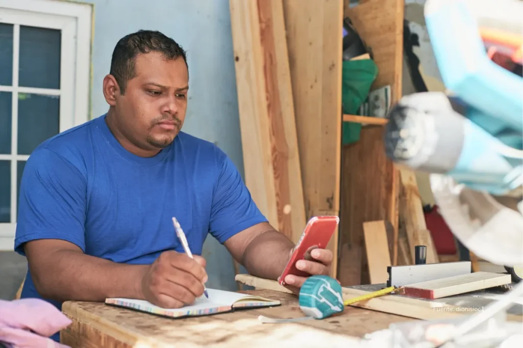 Trabajador manual revisa su teléfono mientras toma notas en una libreta dentro de un taller, con herramientas y materiales de carpintería al fondo.