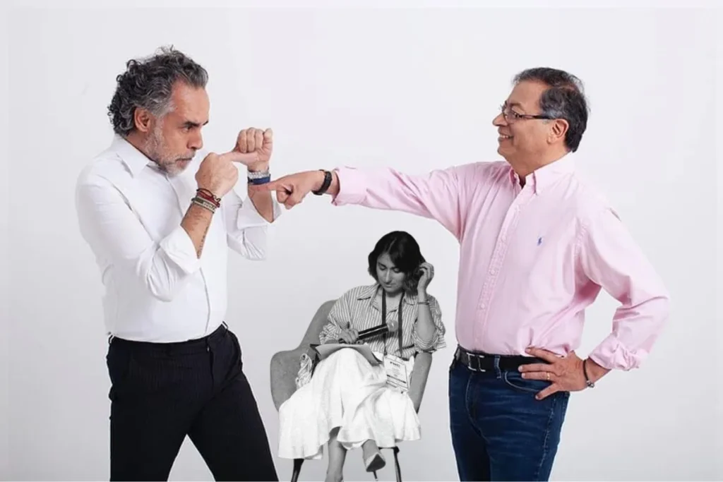 Dos personas interactúan de forma gestual frente a cámara, una en posición de guardia y la otra extendiendo el puño; en el fondo, otra persona sentada escribe en una libreta.