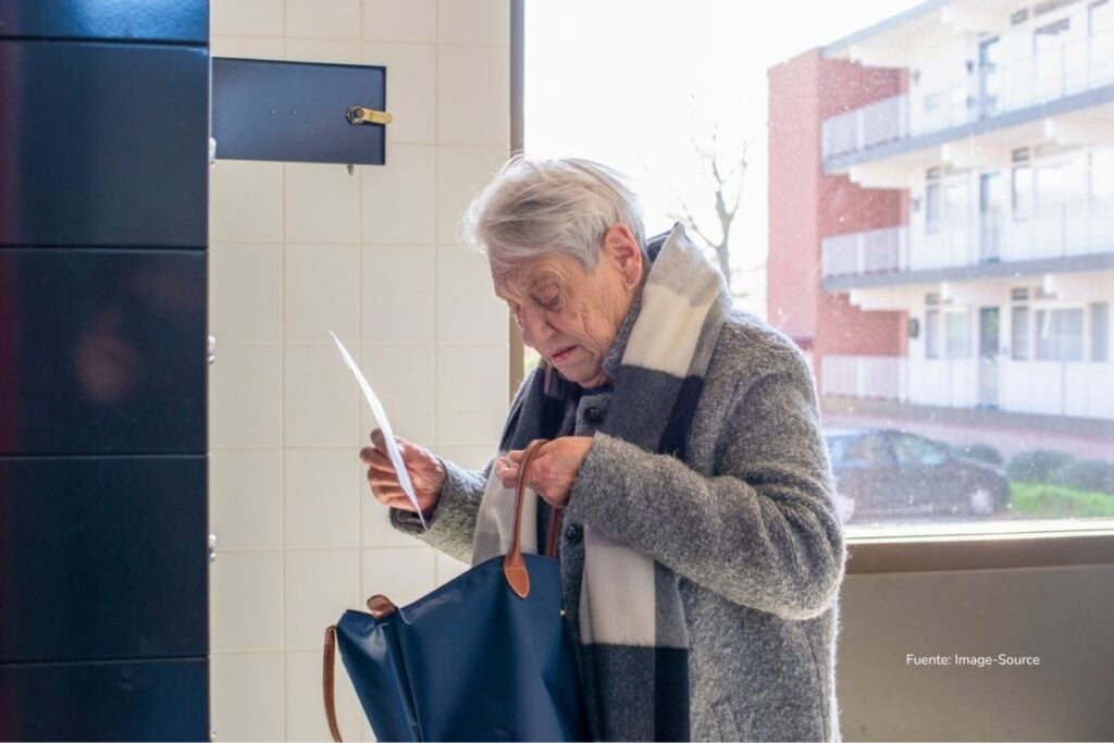 Persona mayor revisa un documento mientras busca algo dentro de su bolso en el pasillo de un edificio residencial. prima de navidad 2025