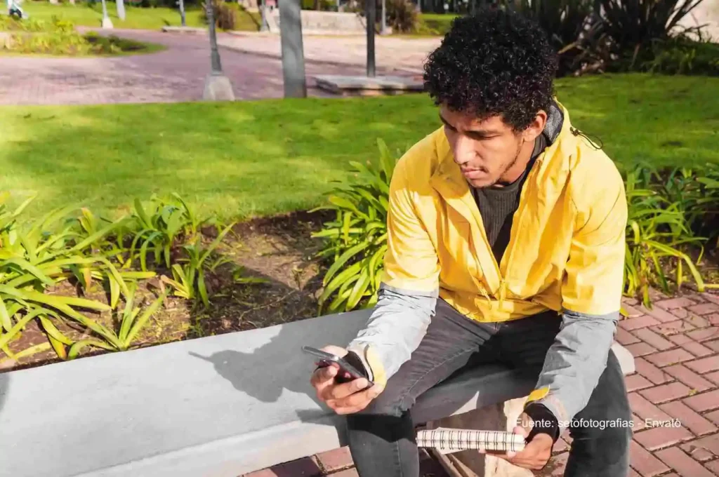 Joven sentado en un parque revisa su celular mientras sostiene un cuaderno sobre las piernas. Integración Tigo Movistar