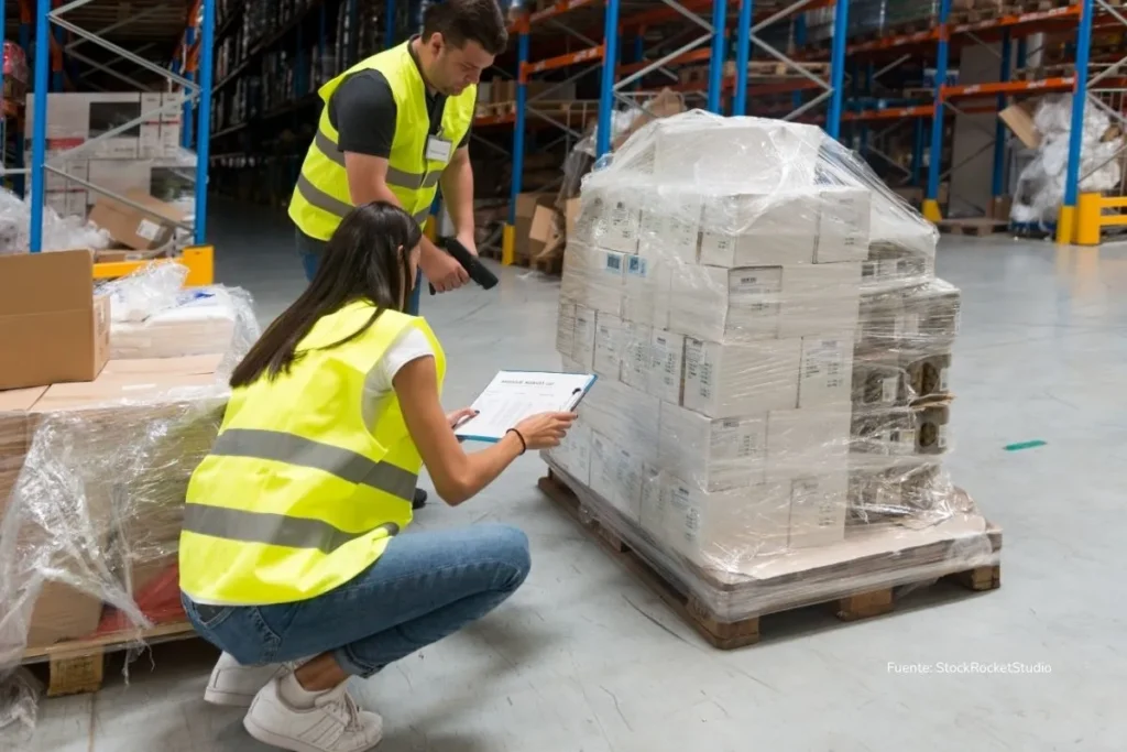 Trabajadores con chalecos reflectivos inspeccionan y registran mercancía empacada en una bodega industrial.