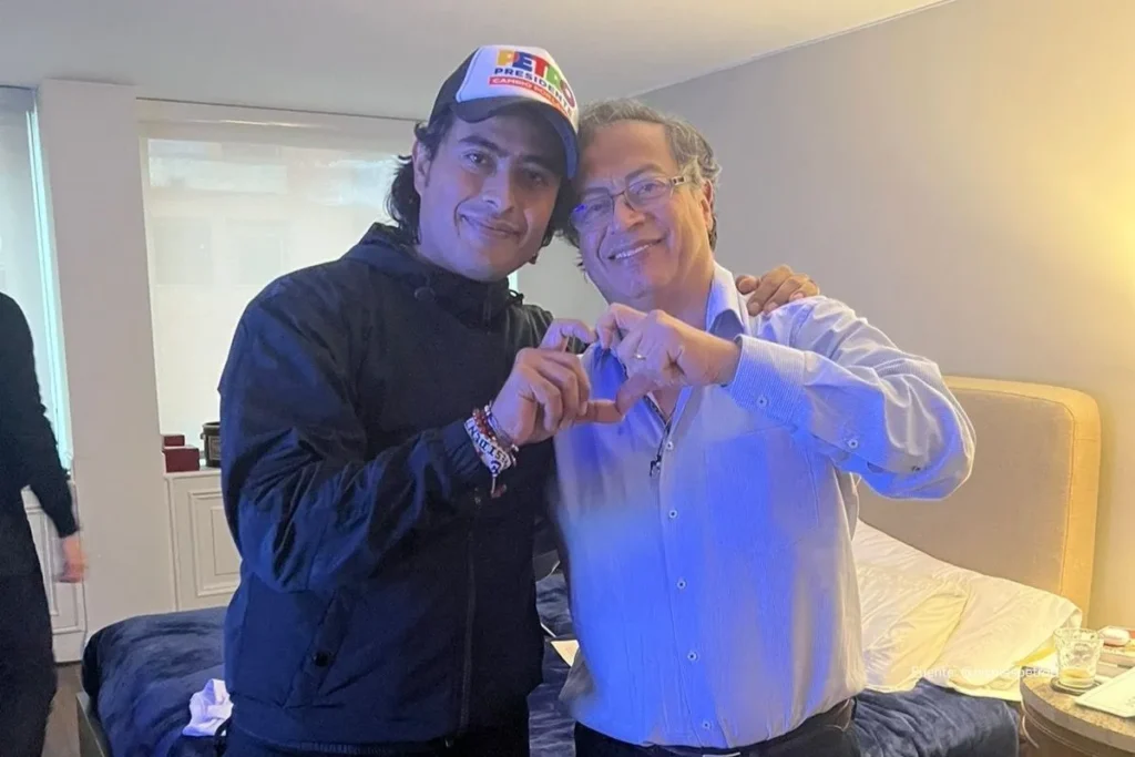 Dos personas posan abrazadas mientras forman un corazón con las manos en el interior de una habitación iluminada.