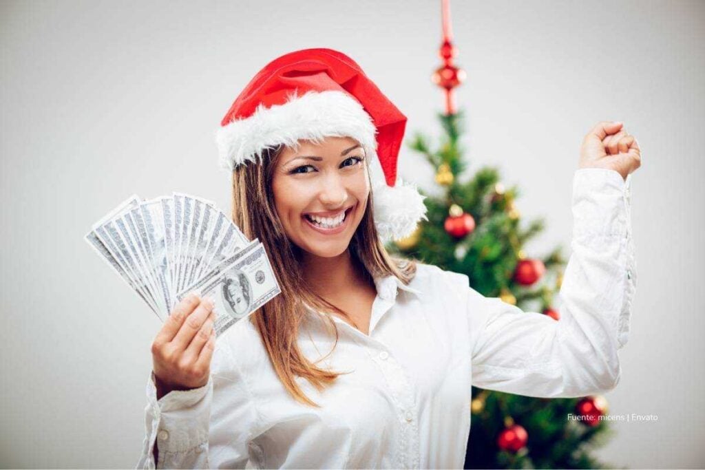 Mujer sonriendo con un gorro de Navidad mientras sostiene varios billetes frente a un árbol navideño decorado.