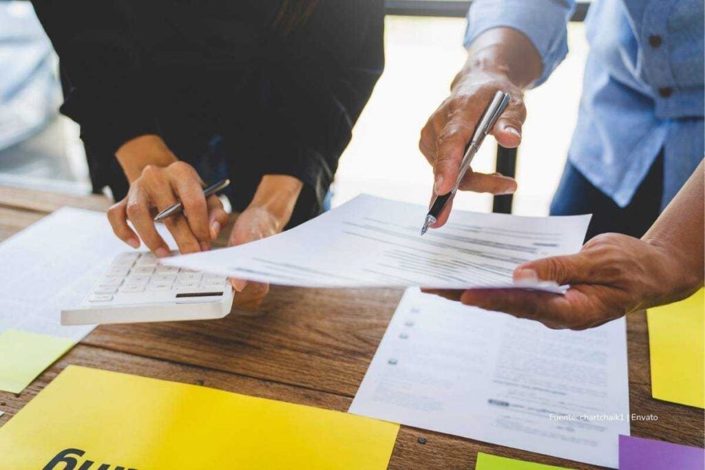 Dos personas revisan documentos impresos mientras una calcula con una calculadora sobre una mesa de trabajo.