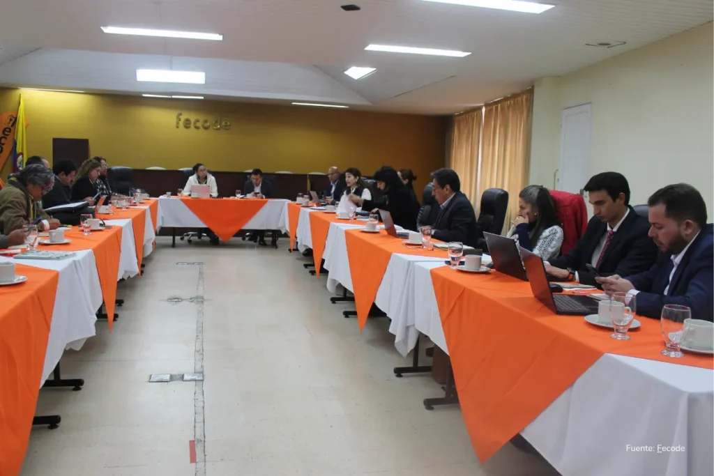 Mesada 14 docentes. Miembros y delegados de Fecode asisten a una asamblea en un salón con tarima, manteles naranjas y banderas del sindicato, mientras un orador interviene frente al auditorio.
