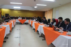 Mesada 14 docentes. Miembros y delegados de Fecode asisten a una asamblea en un salón con tarima, manteles naranjas y banderas del sindicato, mientras un orador interviene frente al auditorio.