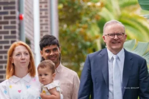 prohibición de las redes sociales en Australia. Un político sonriente camina junto a una pareja y su bebé en un entorno exterior con vegetación, todos mirando hacia adelante.