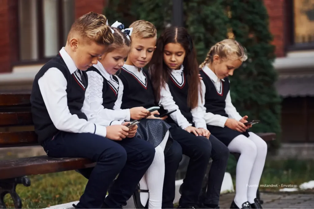 El mundo mira la ley de prohibición de las redes sociales en Australia: Alejará a millones de menores de TikTok e Instagram 2 Cinco niños con uniforme escolar sentados en una banca al aire libre, concentrados en las pantallas de sus teléfonos móviles.