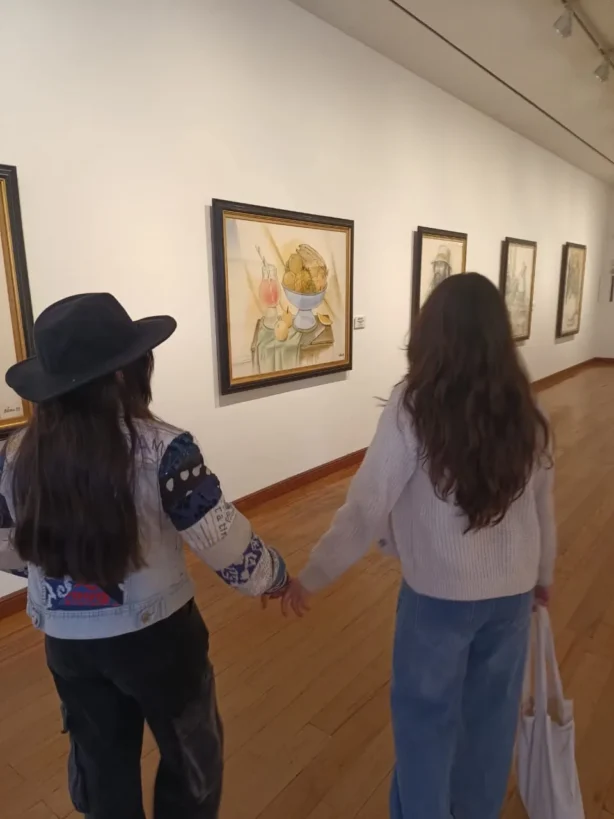 Dos personas de espaldas, tomadas de la mano, recorren una sala de museo y observan pinturas colgadas en una pared blanca.
