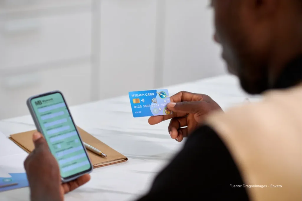 Tarjetas de crédito con cashback en Colombia: cuáles valen la pena y cómo usarlas sin pagar intereses 2 Hombre consulta su teléfono móvil mientras sostiene una tarjeta bancaria en una mesa de trabajo.