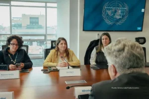 Entrega de medicamentos. Funcionarias participan en una reunión institucional alrededor de una mesa de trabajo, en una sala con pantalla de organismo internacional al fondo.