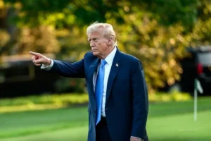 Donald Trump señala con el brazo extendido durante una aparición pública al aire libre, en un entorno ajardinado.