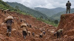 Mineros artesanales cargan sacos y excavan una ladera de tierra rojiza bajo vigilancia armada.