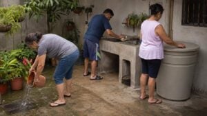 Adultos limpian un lavadero, tapan un depósito y vacían una matera en un patio colombiano.