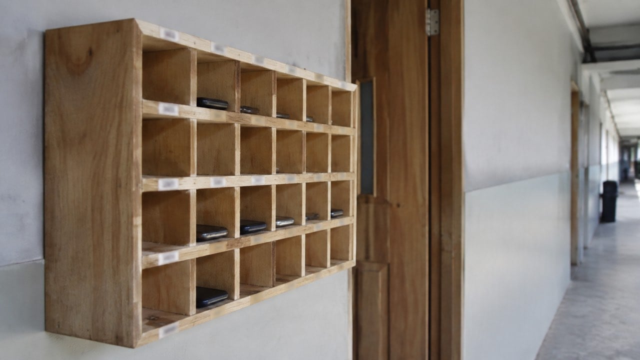 La Secretaría de Educación limita el celular en clases y descansos en Bogotá: la regla cobija a los colegios privados y a docentes 1 Caja de madera con casillas para guardar teléfonos junto a la puerta de un salón de clase