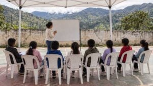 Maestra da clase bajo una carpa a estudiantes sentados en una cancha de pueblo