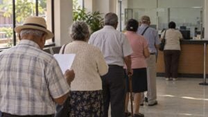 Adultos mayores esperan de espaldas ante una ventanilla; uno usa bastón y otro revisa un papel.