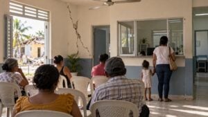 Pacientes esperan en un centro de salud con una grieta en la pared y una madre atiende una ventanilla.