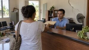 Mujer de espaldas entrega documentos a un asesor en una oficina con una grieta visible en la pared.