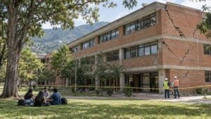Estudiantes conversan frente a un edificio universitario agrietado mientras ingenieros inspeccionan la fachada acordonada.