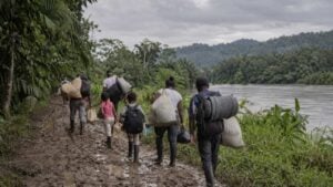 Familias caminan con costales y colchones por un camino embarrado junto a un río en una zona selvática.