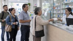 Mujer adulta mayor entrega una fórmula en una droguería mientras otras personas esperan su turno.