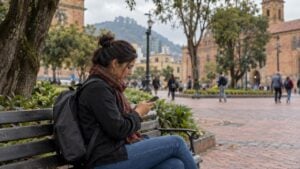 Una mujer revisa ofertas de empleo en su celular en una banca de Bogotá; otras personas caminan al fondo.