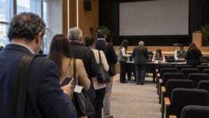 Accionistas adultos hacen fila con documentos y celulares ante mesas de registro en un auditorio.