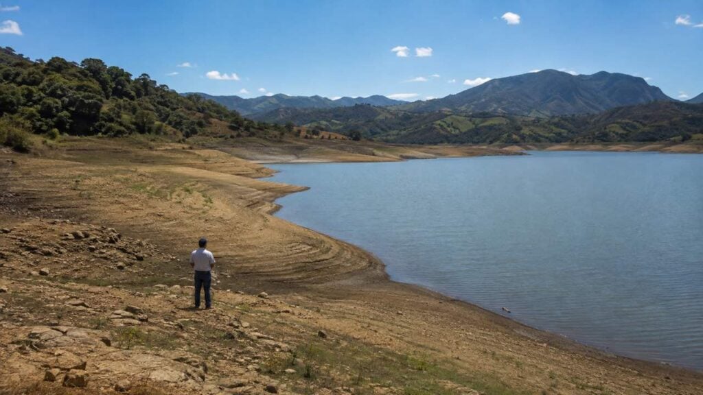 Persona observa un embalse colombiano genérico con nivel bajo visible bajo el sol