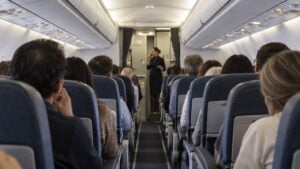 pasajeros en la cabina de un avión comercial miran hacia el pasillo, tensos, mientras una auxiliar de vuelo habla por el intercomunicador.