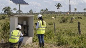 Dos técnicos revisan un sensor sísmico en una caseta con panel solar; al fondo, entre potreros, una torre de perforación.