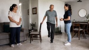 Persona mayor hace un ejercicio de equilibrio acompañada por un fisioterapeuta y un familiar