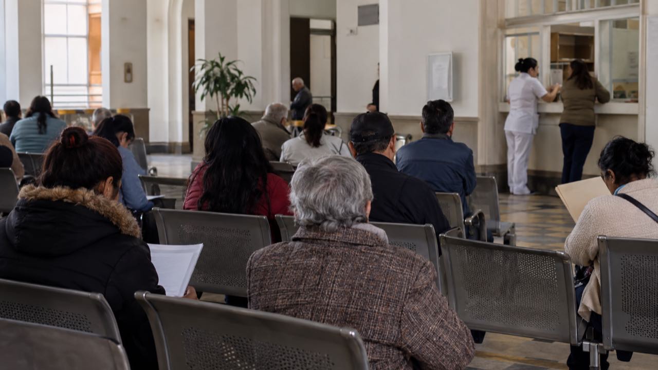 El Hospital de San José dejará de atender a afiliados de Nueva EPS y del Fomag desde el 1 de octubre: las urgencias siguen 1 pacientes esperan sentados en la sala de un hospital de Bogotá mientras una enfermera atiende en una ventanilla.
