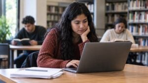 Estudiante universitaria consulta un computador y papeles en una biblioteca con otros estudiantes al fondo.