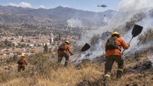 Bomberos forestales combaten un incendio en una ladera seca sobre un pueblo colonial mientras un helicóptero lleva agua al fondo.
