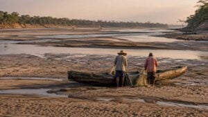 Dos pescadores revisan redes junto a una embarcación varada en la orilla de un río con bancos de arena.