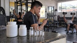 Joven adulto observa una tienda en línea en su celular junto a frascos y ampolletas sin etiqueta en un gimnasio.