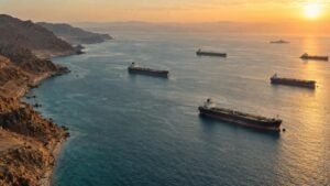 Buques petroleros esperan en un estrecho al atardecer, con costa desértica y un buque de guerra al fondo