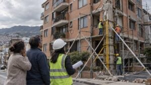 Una ingeniera y dos vecinos miran a obreros que refuerzan con andamios y puntales un edificio de ladrillo agrietado.
