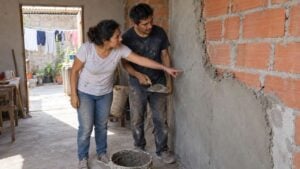 Pareja adulta revisa una pared de ladrillo sin terminar dentro de una vivienda en obra gris.