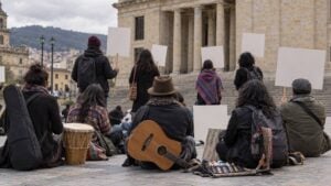 Artistas y gestores culturales de espaldas participan en un plantón pacífico con instrumentos y pancartas en blanco frente a un edificio público genérico de Bogotá.